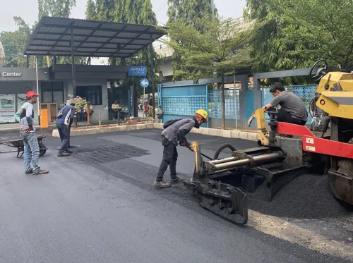Aspal Shell sedang diproses dan diratakan di jalan dengan alat berat, terlihat pekerja memakai helm dan rompi keselamatan.